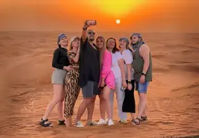 Seven people smiling and posing for a selfie in the desert, with the vibrant colors of the sunset in the background.