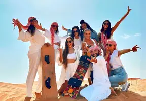 A group of women smiling and posing for a photo with sandboards in the vast expanse of a desert landscape.