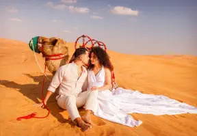 A couple in white outfits sit nd enjoy romantic moments beside a camel in the desert.
