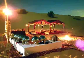 A dinner table set up in the desert at night, illuminated by candlelight and surrounded by sand dunes.